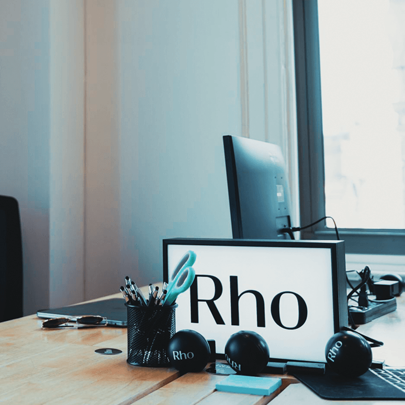 Office desk with a computer, pen holder, scissors, and a lightbox displaying "Rho." Black stress balls are scattered on the wooden surface.